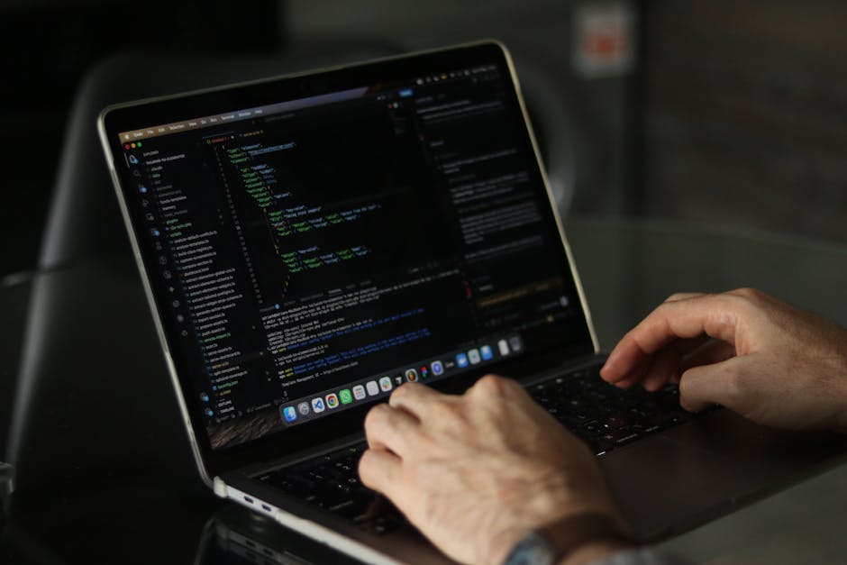 Close-up of a person's hands operating a silver MacBook laptop featuring a dark-themed code editing interface on the screen, situated on a black reflective table. The background is blurred but appears